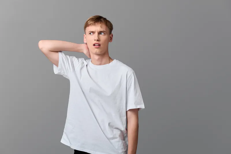 Surprised man in a white t shirt makes a puzzled gesture toward his neck, captured against a neutral backdrop, conveying confusion, hesitation and a moment of uncertainty.