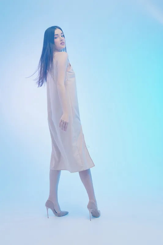 Elegant woman in a beige dress and high heels poses against a soft blue background. The image highlights beauty, fashion, and confident style in a studio setting.