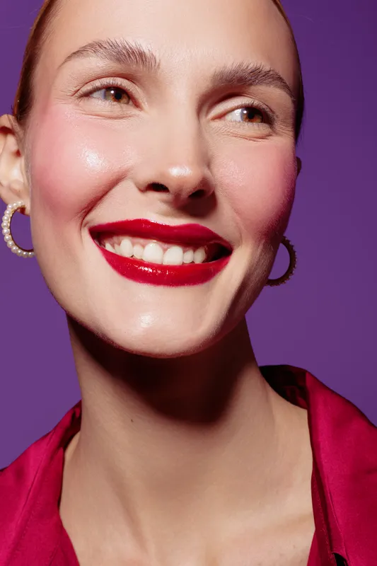 Close-up of smiling woman with red lipstick and hoop earrings on purple background. Concept of beauty, makeup, fashion, and confident female model with natural skin glow.