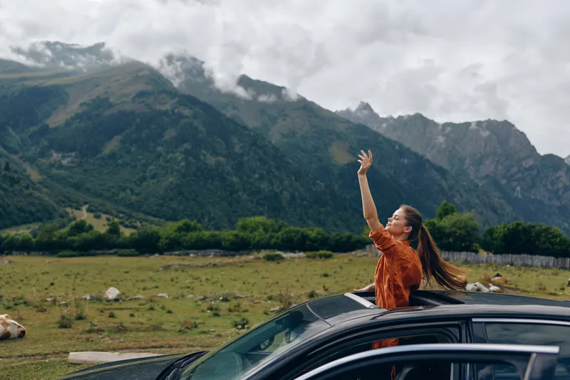 Outdoor woman embraces freedom and adventure from atop a car in a sweeping mountain landscape, arms raised, bright smile, clouds and valley below, conveying travel joy and nature connection