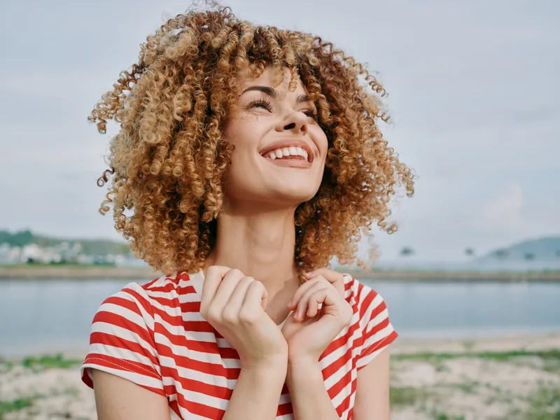 Bright portrait of a joyful woman with curly hair wearing a red and white striped shirt, standing by a calm seaside, smiling with delight and a carefree, luminous vibe.