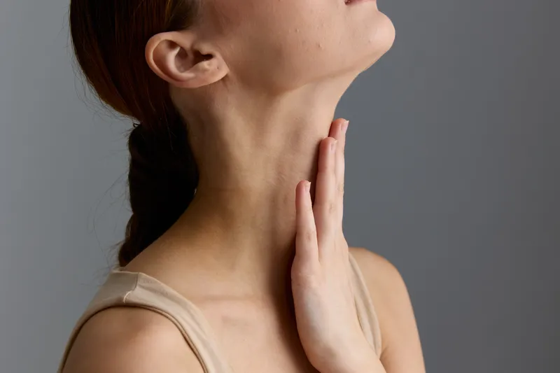 Close-up of a woman touching her neck with a contemplative expression, soft natural lighting, beige tank top, neutral gray background, minimalistic portrait style.