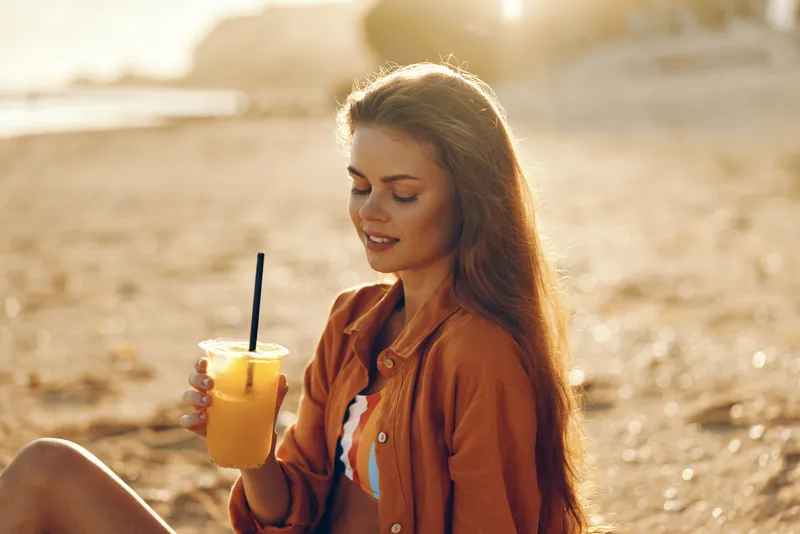 Smiling woman on the beach holding a glass with a straw juice drink at sunset, warm golden sunlight, summer vacation relaxation, wellness mood, calm sea breeze, shallow depth of field soft glow