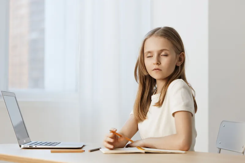 Thoughtful girl with long hair sitting at a desk, surrounded by a bright atmosphere, reflecting on her studies with a laptop nearby