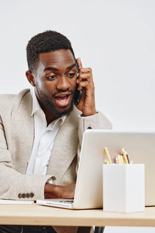 Engaged young African-American man using mobile phone during online work or video call, sitting at desk with laptop and stationery, professional casual attire, isolated on plain white background. Peop