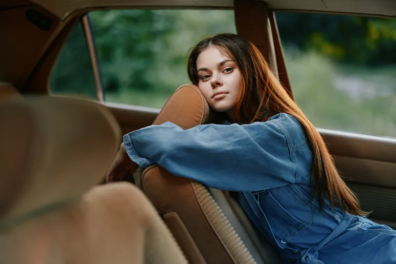 Young woman with long hair in denim jacket relaxing in a vintage car, expressing calmness and nostalgia, surrounded by a lush green background.