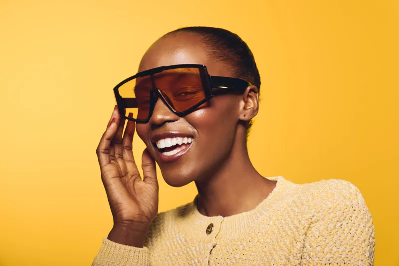 Smiling dark skinned African woman wearing oversized sunglasses and yellow sweater on yellow background. Concept of fashion, beauty, style, and confident mood in studio lighting.