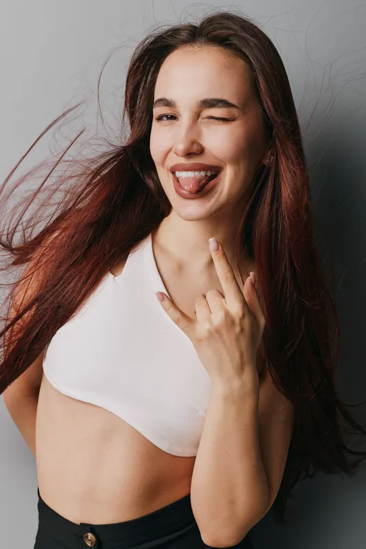 Young woman with long hair making rock sign and winking with tongue out, wearing white crop top and black pants against gray background for fun and attitude portrait.