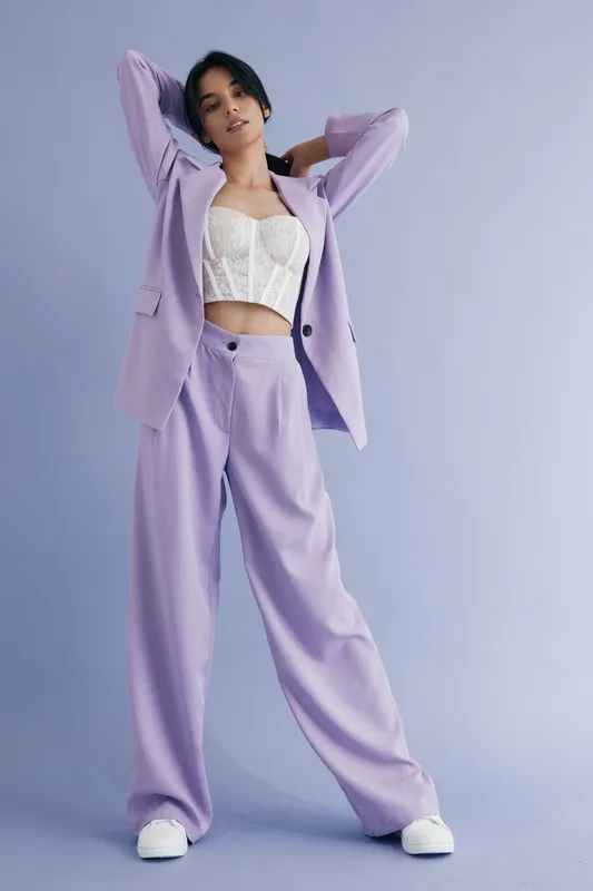 Young woman posing confidently in lavender wide-leg pants and oversized blazer with white corset top, showcasing modern fashion against a pastel background.