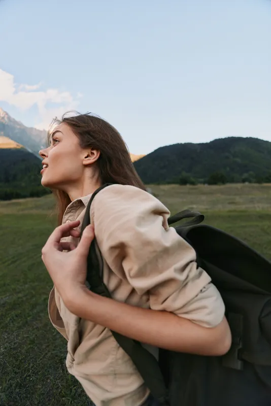 Young woman with backpack in a grassy field with distant mountains, capturing a lifestyle moment in nature, calm and adventurous mood under soft light