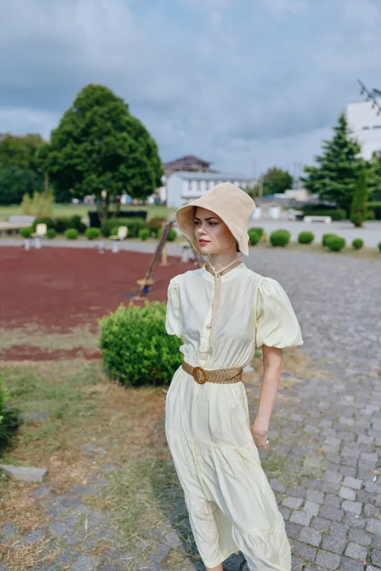 A woman in a light vintage dress and straw hat walks along a cobblestone path in a garden setting, enjoying a calm breezy day that creates a timeless summer scene.