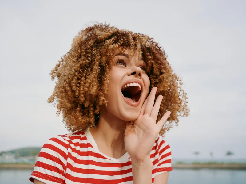 Energetic young woman with curly hair wearing a striped red and white shirt laughs joyfully, mouth open, hand near her face, bright expression against a cloudy sky.