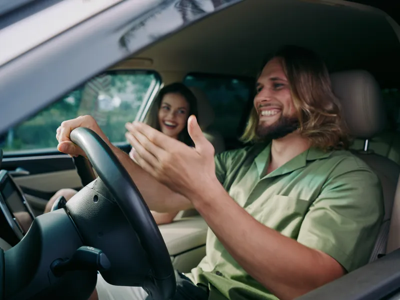Two smiling passengers share a joyful moment inside a modern car, with soft daylight streaming in as the driver gestures happily, capturing a candid, feel good ride.