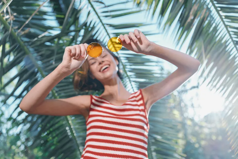 A cheerful woman wearing a striped top smiles while holding sunglasses up, standing beneath bright sunlight and palm fronds, creating a lively, carefree outdoor summer mood.