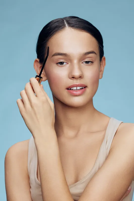 Young woman applying eyebrow gel with a natural makeup look, showcasing beauty routine against a soft blue background featuring a fresh and clean aesthetic