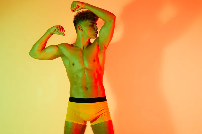 Muscular man posing shirtless in yellow shorts with colorful lighting showing fitness, strength, bodybuilder, and healthy lifestyle in studio environment.