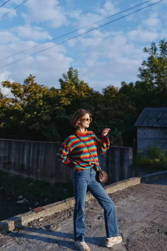 Woman in colorful striped sweater and jeans wearing sunglasses and braided hair, carrying a crossbody bag on a rural road. Standing pose, casual outfit, travel lifestyle and relaxed fashion.