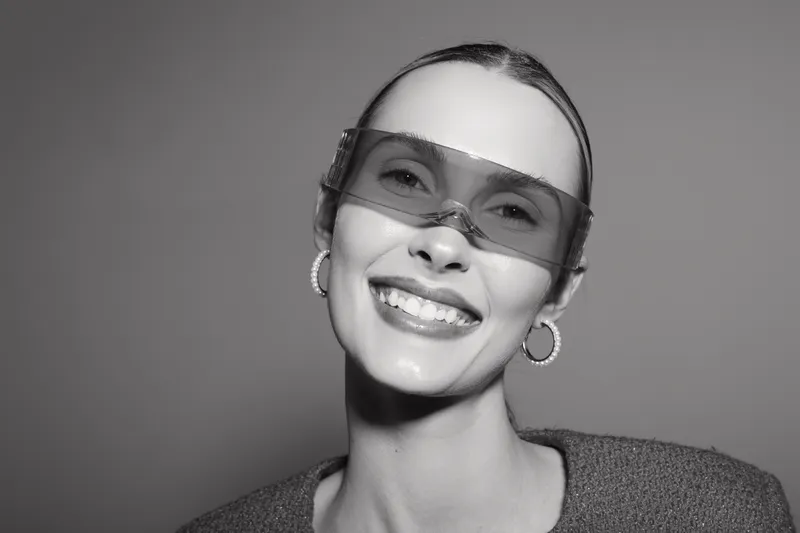 Smiling white woman with light skin wearing transparent sunglasses and hoop earrings on grey background.