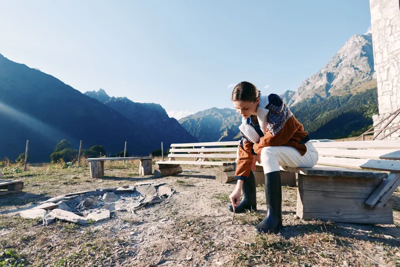 Woman in jacket and boots crouching by a bench in mountains, nature landscape outdoors with clear sky, rural campground scene, exploration and calm morning in scenic alpine view.