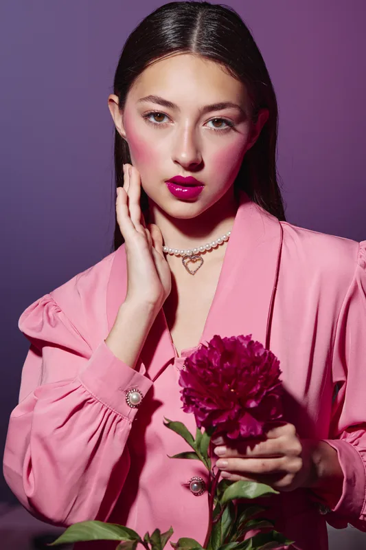 Young fashionable woman wearing a vibrant pink suit, holding a large dark red peony flower. Her makeup features bold pink lips and rosy cheeks. Studio creative beauty shot.