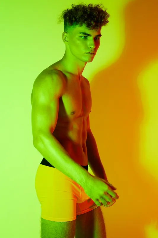Shirtless young man with muscular body wearing bright orange underwear standing against vibrant orange and green background for fitness and summer style portrait.
