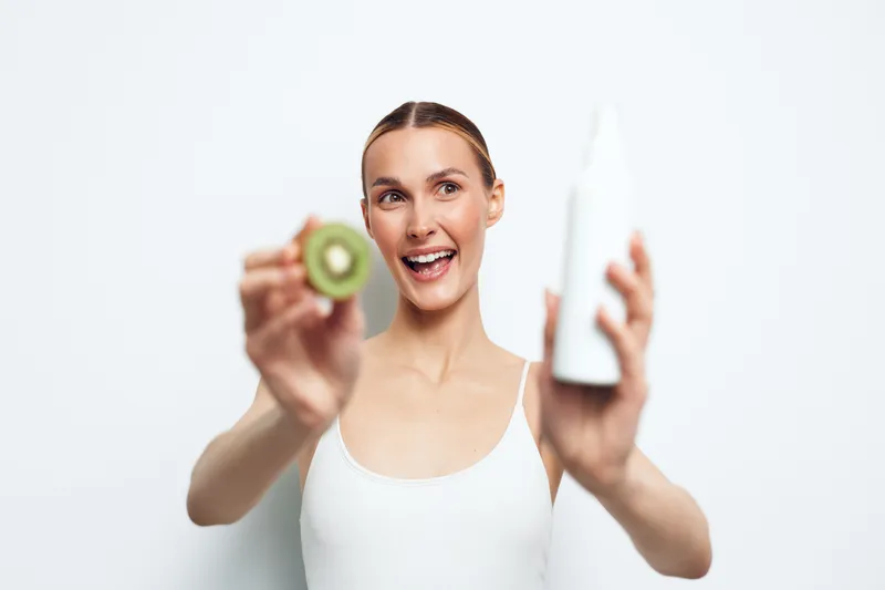 Happy woman with clear skin holding sliced kiwi and white cosmetic bottle, smiling, skincare concept, fresh natural ingredients, bright studio background, health and beauty.