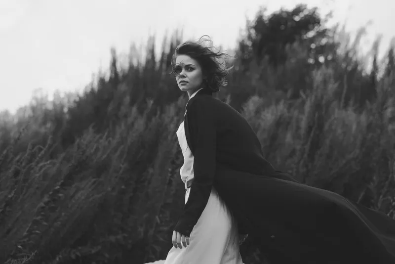 A dramatic monochrome scene of a woman in a flowing coat and dress standing against windy grassland, capturing motion, atmosphere, and resilience in an outdoor fashion moment