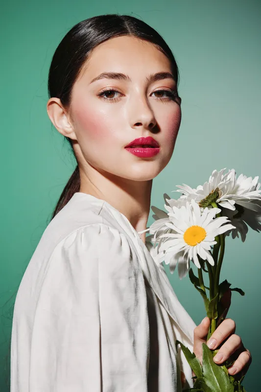 Young woman with vibrant red lipstick holds white daisies against green background in high fashion minimalist beauty portrait. Concept for creative fashion, elegance, and fresh spring style.