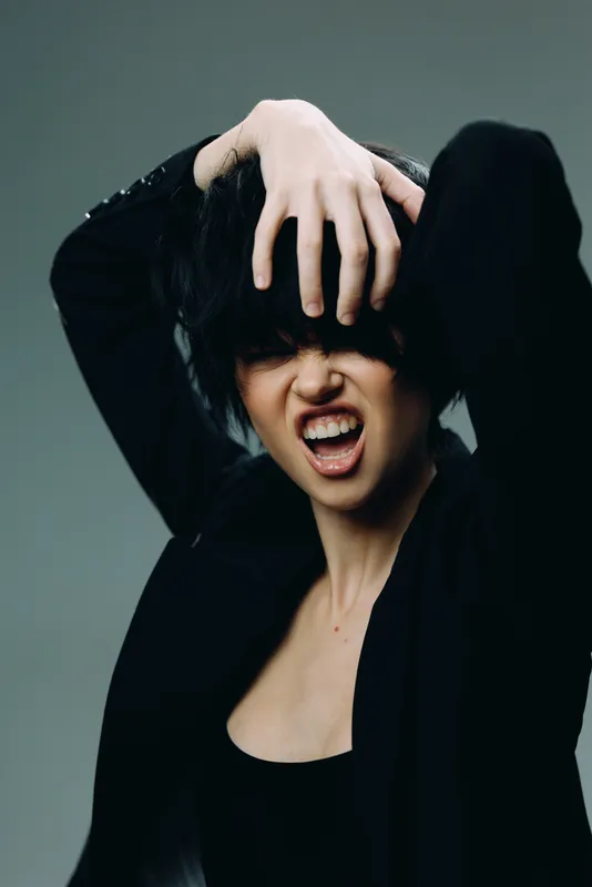 expressive woman with messy hair showing intense emotion in dark casual outfit against neutral background