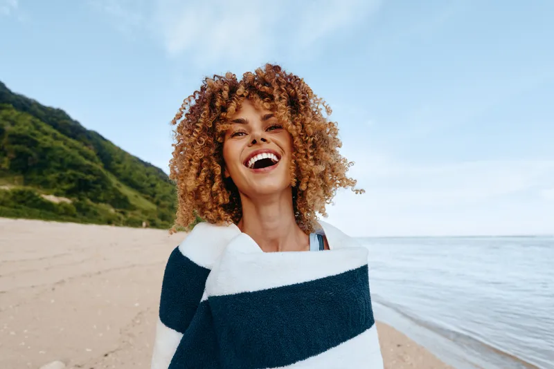 Woman, curly hair, smiling, laughter on beach, sea waves behind, outdoors in summer, vacation happiness, natural light, coastal breeze