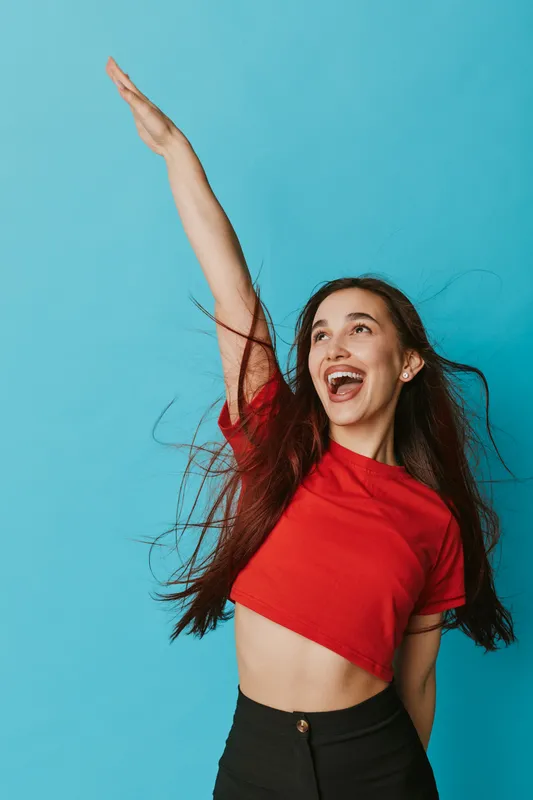 Young woman with long hair wearing red crop top and black pants smiling and raising arm against blue background. Happiness, joy, casual style and positive energy in studio portrait.