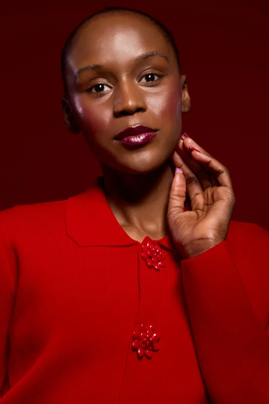 Portrait of a dark skinned African woman with glowing skin wearing a red sweater, posing with her hand near face. Studio lighting highlights her smooth complexion and elegant expression.