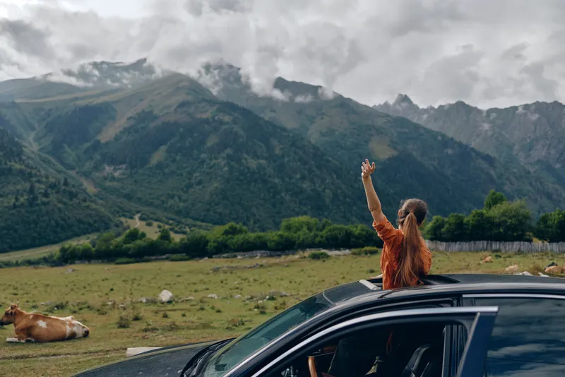 Traveler raises an arm from a car roof as mountains and fields stretch into the distance creating an outdoor adventure atmosphere and a sense of freedom in a wide natural landscape
