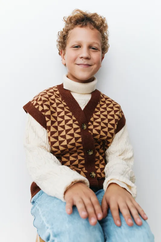 Authentic portrait of a cheerful child wearing a geometric knitted vest over a cozy sweater and jeans, capturing a candid school day moment with a warm, playful game mood.