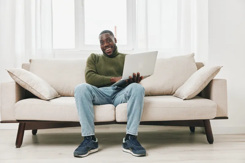 Happy African American man working on a laptop at home, wearing a green sweater and jeans, sitting on a cozy sofa in a bright, minimalist living room