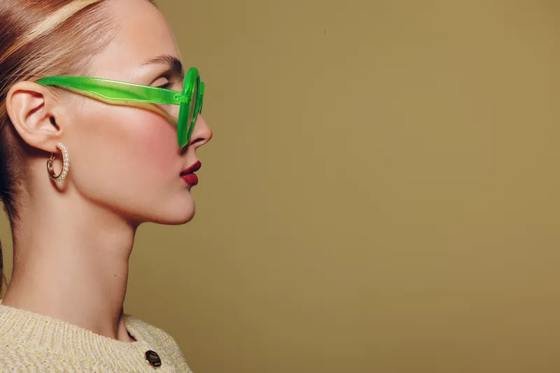 Profile of a stylish woman wearing green transparent sunglasses and gold hoop earrings against a beige background. Concept of fashion, accessories, and modern style.