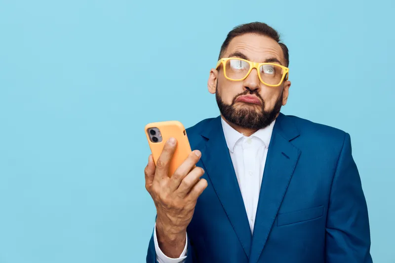 A business professional man stands against a solid colored background, expressing a puzzled emotion while checking a mobile device, creating a bold corporate portrait with a quirky vibe.
