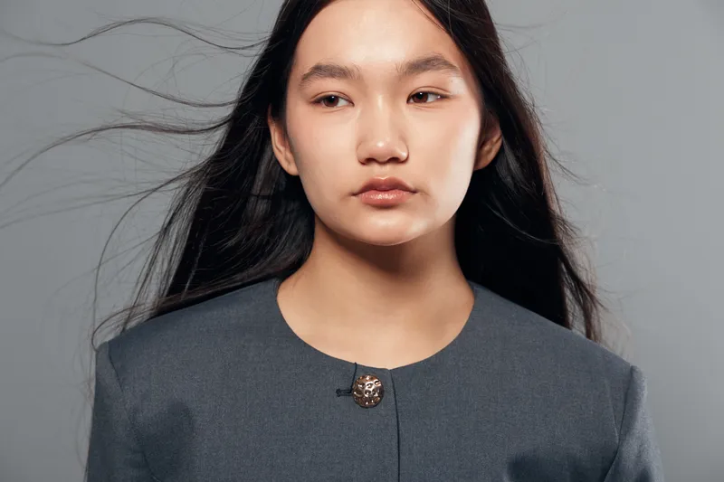 Young Asian woman with long black hair wearing gray jacket looking away on gray background. Studio portrait of confident female with natural makeup and serious expression with loose hair.