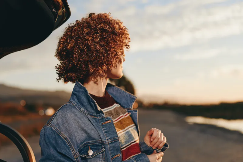 Woman and child in denim outdoors at sunset, curly hair close up, holding hands while walking near coast, warm golden hour lifestyle, candid family moment, soft breeze and calm mood