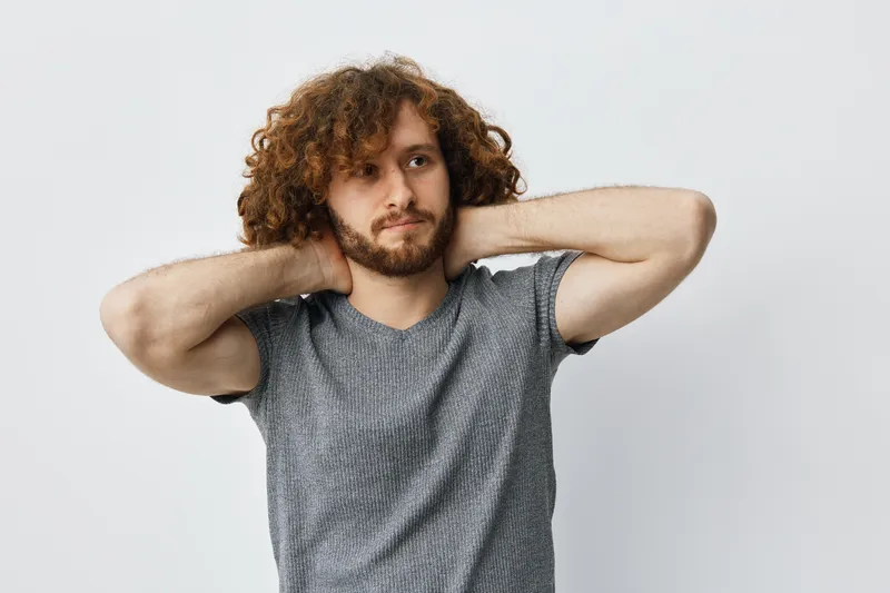 Young man with curly hair in a casual gray shirt, expressing calmness and relaxation against a light background, ideal for lifestyle and wellness themes
