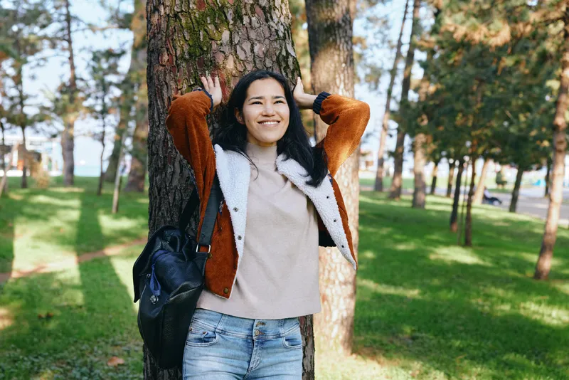 Woman leaning against a tree in a sunlit park, smiling, wearing a brown jacket and sweater, casual denim, backpack on shoulder, enjoying nature and fresh air during a relaxed outdoor moment