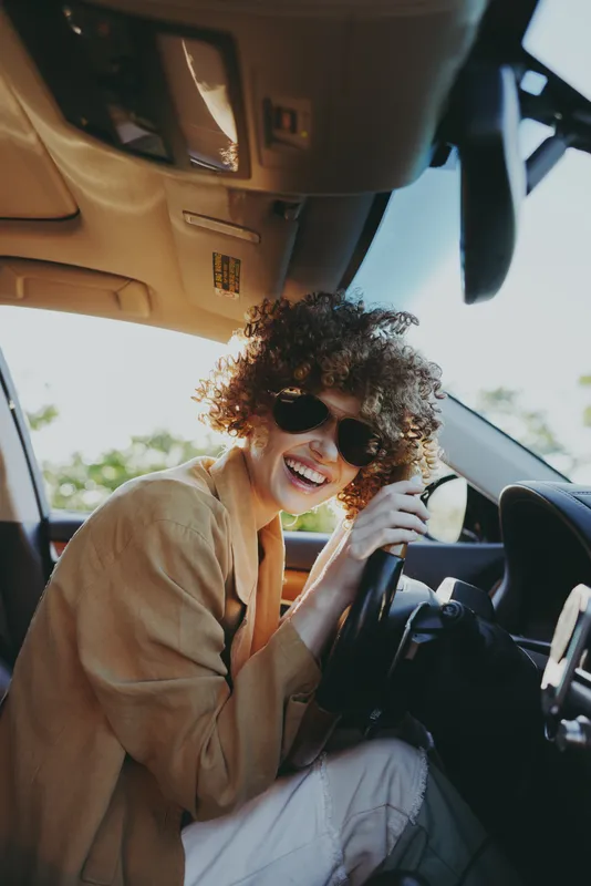 A stylish woman driver wears sunglasses and a beige jacket, smiling while steering in a sunny car interior. A candid lifestyle moment with retro film color and warm tones, conveying confident