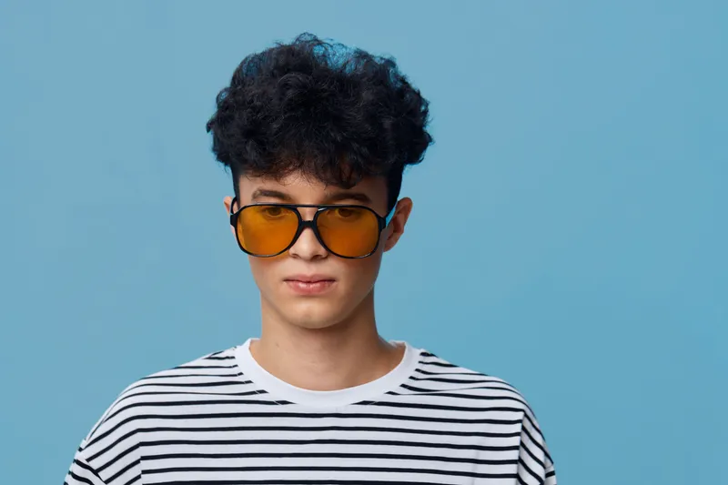 Young man with curly hair wearing oversized amber sunglasses and a white striped shirt standing against bright blue background, showcasing casual modern style.