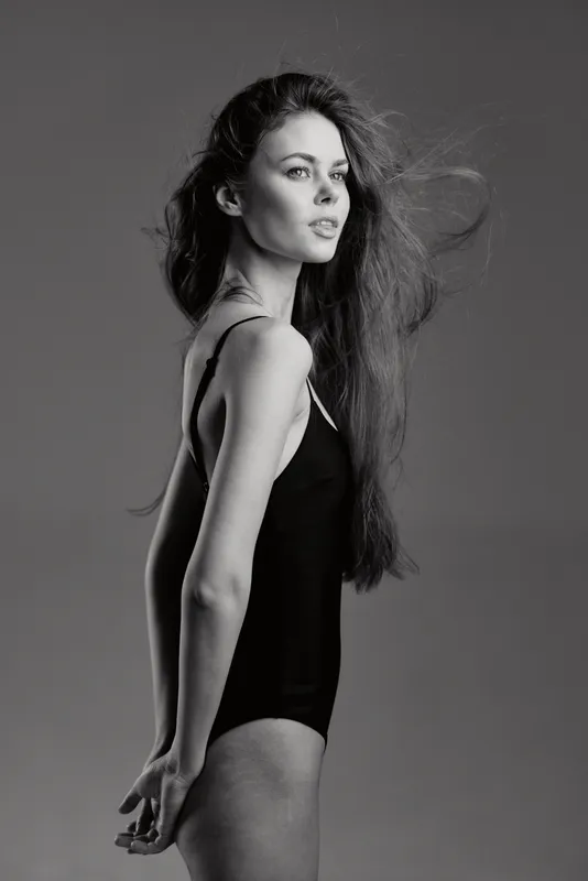 Black and white portrait of a thoughtful woman in a black bodysuit with flowing hair on a plain background.
