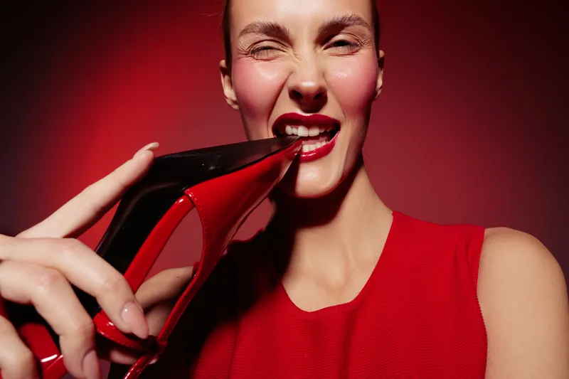 Young woman in red dress bites red high heel shoe with playful expression on red background. Concept of fashion, bold style, attitude, and creative portrait photography.