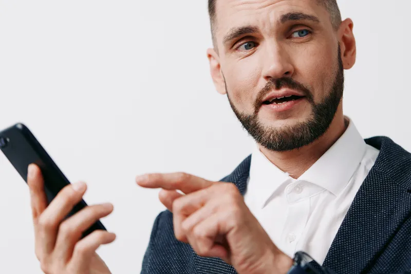 A professional businessman appears focused while using a smartphone, expressing curiosity and engagement. The subject stands against an isolated colored background, showcasing confidence, drive, and