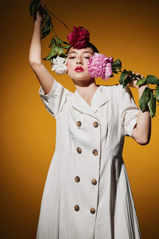 Stylish woman in white dress poses with colorful flowers herself against warm orange background. Creative beauty and fashion concept with bold makeup and artistic composition.