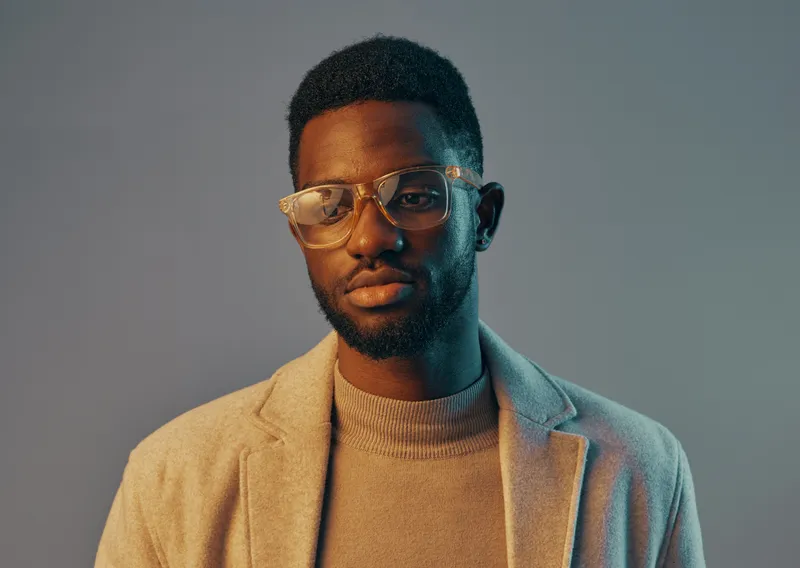 Stylish young Black man in glasses wearing a beige blazer and turtleneck, showcasing confidence against a soft gradient background emphasizing fashion and modern aesthetics