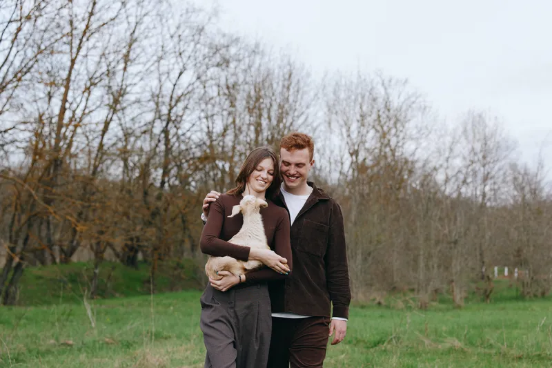 Happy couple outdoors with their dog, embracing in a grassy field, smiling and affectionate, casual wear, warm mood, natural light, candid lifestyle moment, serene countryside atmosphere.
