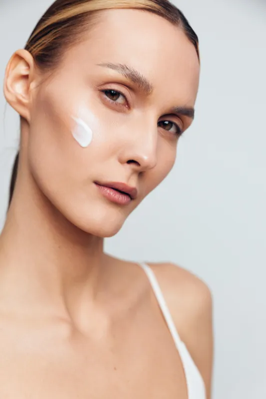 Closeup portrait of young woman with natural skin applying cream on cheek, soft lighting, neutral background, skincare routine, fresh and minimal beauty concept.